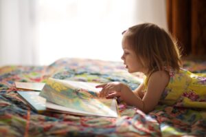 A little girl in a yellow dress reads a picture book on her bed.