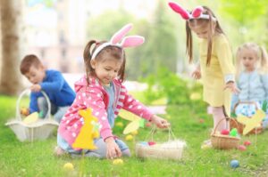 A group of children in bright clothes and wearing bunny ears hunt for colourful Easter eggs in a meadow.