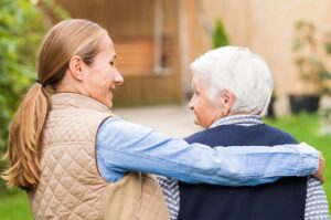 A caring woman puts her arm around an elderly lady as they walk outdoors.