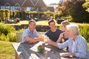A multi-generational family raise a glass while sitting on a bench in a beer garden in the sun.