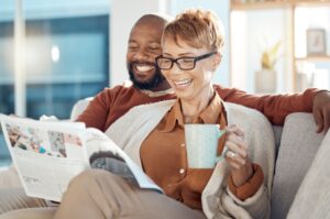 A couple sitting on a sofa laugh as they drink tea and read a newspaper together.