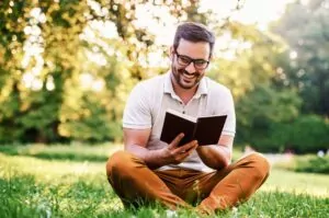 A man sits cross-legged in a field smiling as he reads a book.