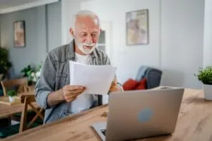 A man sits at a table reading his pension statement, with his laptop in front of him.